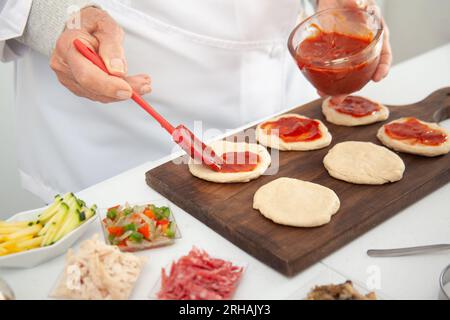 Femme senior ajoutant de la sauce pizza à la pâte étirée. Délicieuse préparation de mini pizzas maison. Banque D'Images