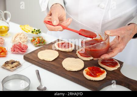 Femme senior ajoutant de la sauce pizza à la pâte étirée. Délicieuse préparation de mini pizzas maison. Banque D'Images