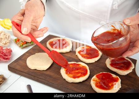 Femme senior ajoutant de la sauce pizza à la pâte étirée. Délicieuse préparation de mini pizzas maison. Banque D'Images