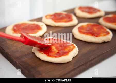 Femme senior ajoutant de la sauce pizza à la pâte étirée. Délicieuse préparation de mini pizzas maison. Banque D'Images