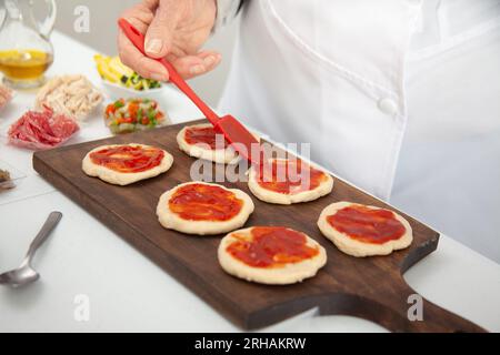 Femme senior ajoutant de la sauce pizza à la pâte étirée. Délicieuse préparation de mini pizzas maison. Banque D'Images