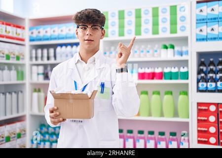 Adolescent hispanique travaillant à la pharmacie pharmacie tenant la boîte avec des pilules souriant heureux pointant avec la main et le doigt sur le côté Banque D'Images