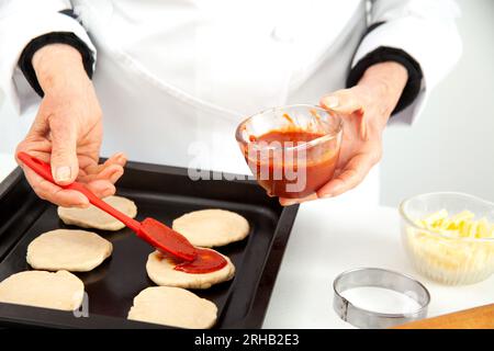 Femme senior ajoutant de la sauce pizza à la pâte étirée. Délicieuse préparation de mini pizzas maison. Banque D'Images