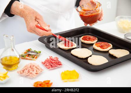 Femme senior ajoutant de la sauce pizza à la pâte étirée. Délicieuse préparation de mini pizzas maison. Banque D'Images