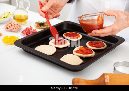 Femme senior ajoutant de la sauce pizza à la pâte étirée. Délicieuse préparation de mini pizzas maison. Banque D'Images