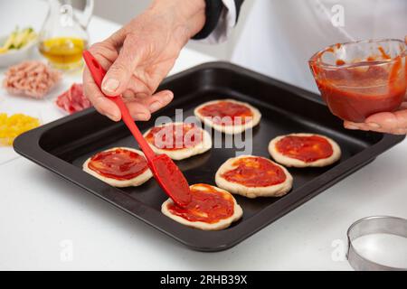 Femme senior ajoutant de la sauce pizza à la pâte étirée. Délicieuse préparation de mini pizzas maison. Banque D'Images