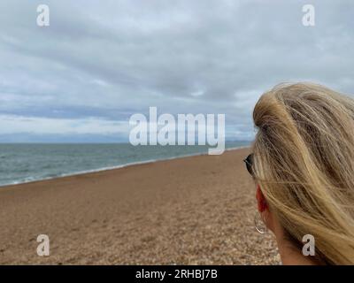 Vue arrière d'une femme assise sur Chesil Beach regardant la mer en été, Dorset, Angleterre, Royaume-Uni Banque D'Images