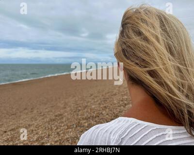 Vue arrière d'une femme assise sur Chesil Beach regardant la mer en été, Dorset, Angleterre, Royaume-Uni Banque D'Images