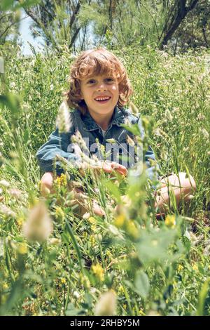 Joyeux garçon mignon prèteen aux cheveux bouclés assis avec les jambes croisées dans le champ de floraison vert dans la journée ensoleillée d'été Banque D'Images