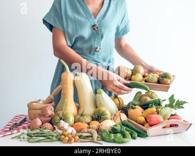 Cultivez la femelle méconnaissable en robe debout près d'une pile de citrouilles fraîches et de poivrons et d'oignons avec l'ail et les pois placés sur la table et tenant le tomat Banque D'Images