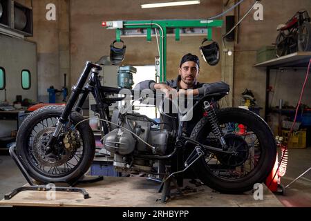 Mécanicien barbu sérieux en uniforme, appuyé sur une moto démontée et regardant la caméra dans un atelier minable Banque D'Images