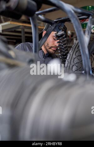 Du dessous du technicien masculin hispanique concentré en uniforme et chapeau fixant la moto pendant la journée de travail dans l'atelier de réparation Banque D'Images