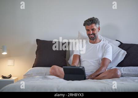 Entrepreneur masculin mature en vêtements de nuit souriant et regardant l'écran de la tablette tout en travaillant sur le projet à distance dans la chambre à coucher à la maison Banque D'Images