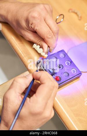 D'en haut les mains de la récolte maître méconnaissable peinture dentier blanc avec brosse tout en étant assis à table dans le laboratoire dentaire moderne Banque D'Images