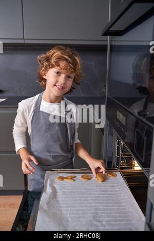 Enfant souriant en tablier gris mettant des biscuits sur du papier sulfurisé se préparant à les cuire dans la cuisinière et regardant la caméra Banque D'Images