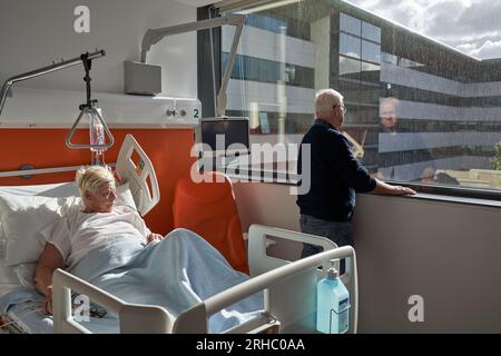 Vue latérale d'un homme âgé contrarié avec de courts cheveux blancs regardant par la fenêtre alors qu'il se tenait à l'hôpital près d'une femme malade couchée sur le canapé pendant la réhabilitation Banque D'Images