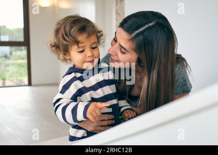 Femme souriante avec de longs cheveux foncés embrassant mignon enfant aux cheveux bouclés dans un pull rayé et parlant amicalement dans une pièce spacieuse et lumineuse Banque D'Images