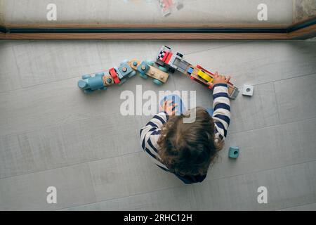 Vue de dessus d'un enfant anonyme aux cheveux bouclés assis sur le sol près de la fenêtre et jouant avec des voitures jouets et le train Banque D'Images