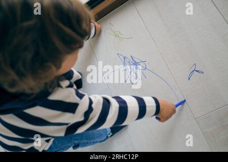 D'en haut de tout-petit aux cheveux bouclés anonyme en dessin de vêtements décontractés avec des marqueurs colorés sur le sol stratifié près de la fenêtre Banque D'Images