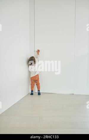 Vue arrière d'un petit garçon anonyme avec les cheveux bouclés debout sur la pointe des pieds tout en ouvrant la porte blanche dans le salon à la maison Banque D'Images