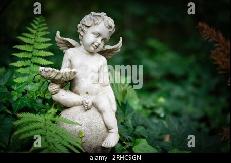 petit putto avec des ailes et un bol à la main assis sur une boule sur une tombe entourée de fougères Banque D'Images