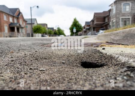 Nid de poule sur la rue résidentielle - ciel couvert et humeur sombre - trou circulaire et profond - maisons et arbres en arrière-plan Banque D'Images