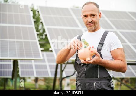 Investisseur mettant des euros à la tirelire sur fond de panneaux solaires. Homme compétent investissant dans l'énergie verte pour le profit futur. Homme adulte en salopette heureux de l'investissement qu'il a fait. Banque D'Images