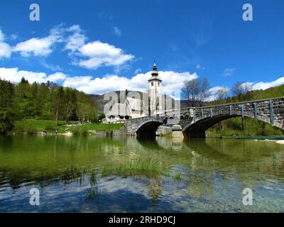 Belle vue sur le St. Jean l'église baptiste à côté du lac Bohinj à côté d'un pont en pierre voûté et un reflet dans le lac et des nuages blancs en b. Banque D'Images