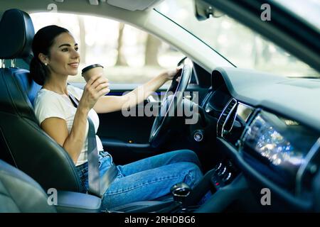 Heureuse jeune femme avec du café pour aller conduire sa voiture. Vue latérale de la femme conduisant la voiture avec le café pour aller à la main. Jeune femme buvant du café pendant qu'elle drivi Banque D'Images