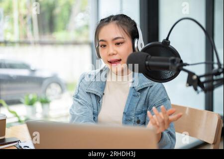 Une animatrice de radio ou podcasteuse asiatique professionnelle et focalisée regarde son écran d'ordinateur portable, parle dans un micro, annonce des nouvelles et large Banque D'Images