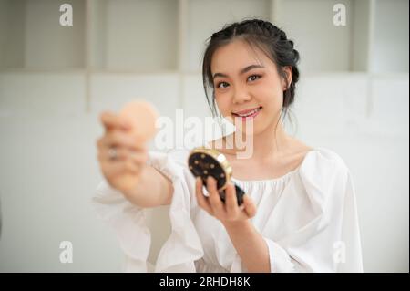 Une attrayante influenceuse de beauté asiatique appliquant son maquillage, montrant son nouveau produit cosmétique à une caméra et créant sa vidéo tutoriel de maquillage Banque D'Images