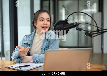 Une belle et professionnelle animatrice de radio asiatique parle dans un micro, annonçant ou partageant des nouvelles intéressantes avec son public en ligne pendant Banque D'Images