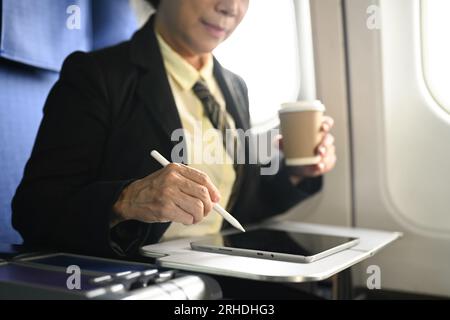 Femme d'affaires mature assise à son siège dans l'avion et utilisant la tablette numérique. Banque D'Images