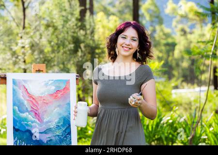 Heureux jeune artiste australien tenant un pot de peinture blanche et un pinceau à côté d'œuvres d'art. Banque D'Images