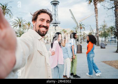 Beau jeune homme adulte prenant un portrait selfie tenant la main de sa petite amie alors qu'ils marchent dans la ville avec leurs amis joyeux. Les gens internationaux s'amusent ensemble le week-end. Photo de haute qualité Banque D'Images