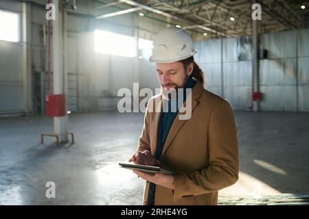 Jeune entrepreneur confiant ou propriétaire d'entrepôt défilant en tablette à travers des informations en ligne ou des documents avec des croquis techniques Banque D'Images