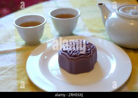 Une pâte Mooncake chinoise violette embossée de fleurs sur une assiette blanche avec deux tasses à thé chinois et une théière sur une nappe à carreaux de citron Banque D'Images