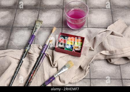 Peintures d'artiste avec des pinceaux, tissu et verre sur fond de carreaux gris Banque D'Images