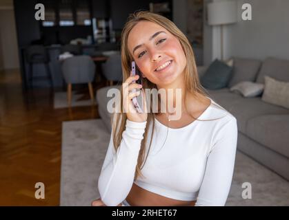 Femme millénaire souriante parlant au téléphone à la maison, jeune fille heureuse tient le téléphone portable faisant l'appel de réponse, adolescent attrayant ayant plaisant con Banque D'Images