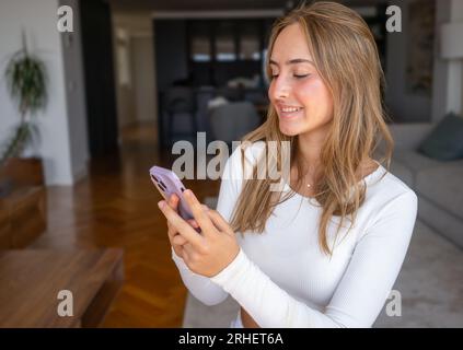 Femme millénaire souriante parlant au téléphone à la maison, jeune fille heureuse tient le téléphone portable faisant l'appel de réponse, adolescent attrayant ayant plaisant con Banque D'Images