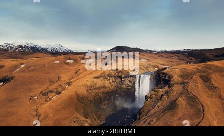 Vue aérienne de la cascade arctique de skgafoss qui descend des collines gelées, créant des paysages islandais spectaculaires. Cascade scandinave massive tombant des falaises brunes. Ralenti. Banque D'Images