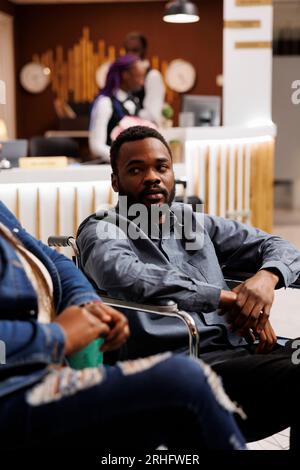 Jeune homme afro-américain touriste en fauteuil roulant arrivant à l'hôtel pour handicapés, assis dans le hall parlant avec sa petite amie ou sa femme en attendant la procédure d'enregistrement. Utilisation d'un fauteuil roulant pendant le voyage Banque D'Images