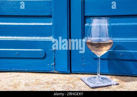 Vin rose sur un balcon en pierre avec volets bleus colorés et la mer Égée bleue au loin : l'île de Tinos Grèce Banque D'Images