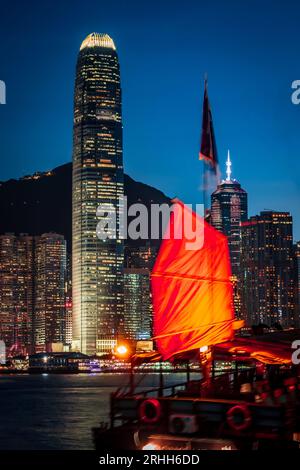 Superbe vue nocturne d'une jonque brillamment illuminée dans le port Victoria en face de l'horizon fantastique de Hong Kong avec ses impressionnants gratte-ciel Banque D'Images