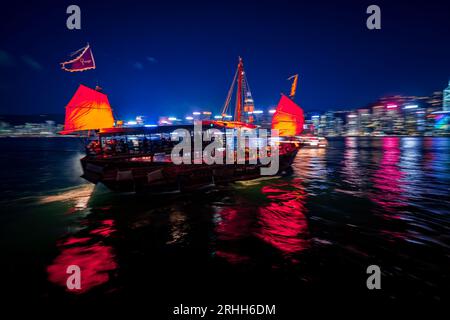 Superbe vue nocturne d'une jonque brillamment illuminée dans le port Victoria en face de l'horizon fantastique de Hong Kong avec ses impressionnants gratte-ciel Banque D'Images