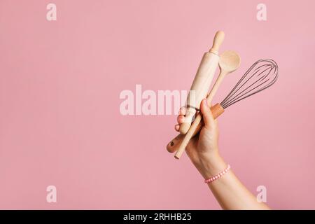 Main féminine tenant des ustensiles de cuisine pour la nourriture et la boulangerie sur fond rose. fouet, rouleau à pâtisserie et cuillère en bois Banque D'Images