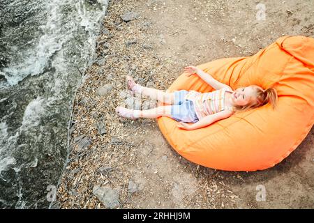 Mignon bébé fille couchée sur une chaise à sac près de la rivière. Petit enfant actif jouant à l'aire de jeux de la cour de l'école. Les enfants jouent et grimpent à l'extérieur sous le soleil Banque D'Images
