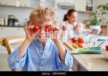 Heureux garçon blond couvrant les yeux avec des tomates cerises rouges dans la cuisine à la maison Banque D'Images