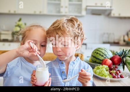 Fille mangeant parfait à l'aide de cuillère avec frère tout en étant assis par des fruits sur la table dans la cuisine à la maison Banque D'Images