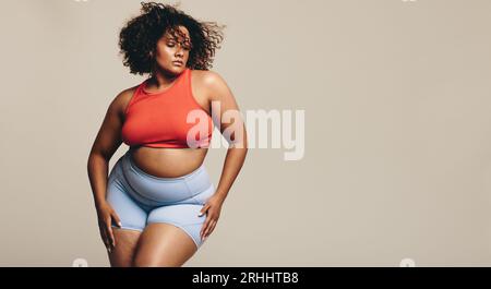 Femme sûre d'elle-même frappe une pose confiante dans des vêtements de sport dans un studio, montrant son physique tonique. Jeune femme athlétique embrassant son séjour Banque D'Images